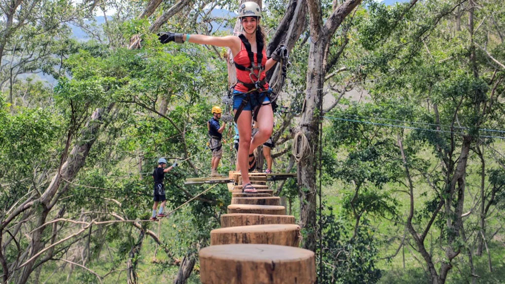 Woman wearing a helmet and safety harness balancing on a suspended wooden log bridge high above a forested ravine near Puerto Vallarta.