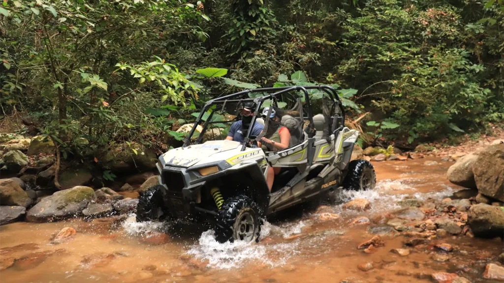 Polaris RZR UTV carrying helmeted riders through a shallow rocky stream surrounded by tropical forest during the All-Terrain Safari in Puerto Vallarta.