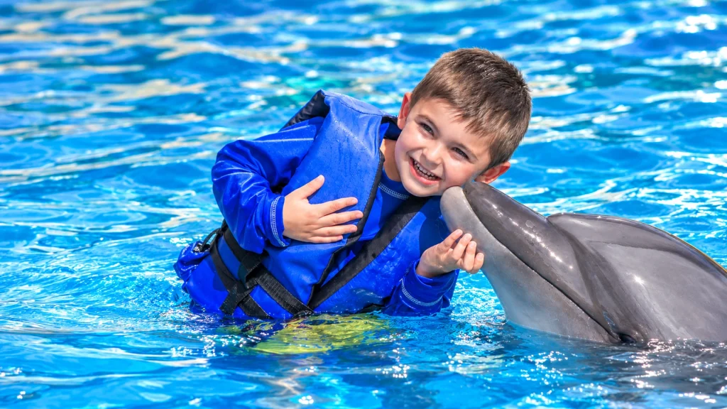 Smiling young boy in a blue life jacket receiving a kiss from a bottlenose dolphin during Dolphin Kids in Nuevo Vallarta.