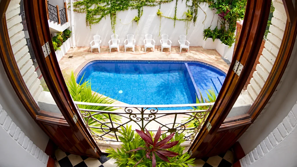 Courtyard swimming pool at Hacienda de Vallarta Centro, viewed through open wooden doors with tropical plants and poolside chairs.