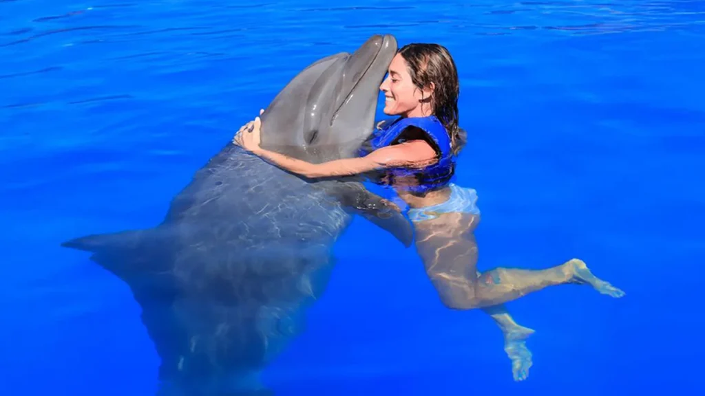 Woman in a blue life jacket embracing a bottlenose dolphin in a bright blue swimming pool.