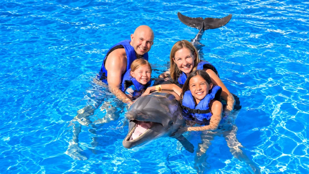 Family of four wearing blue life jackets smiling beside a bottlenose dolphin in a turquoise pool during a private dolphin swim in Nuevo Vallarta.