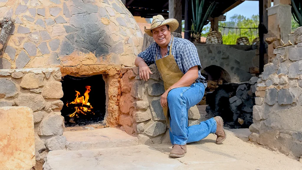 Smiling man wearing a cowboy hat and leather apron beside a traditional stone wood-fired oven with flames inside.