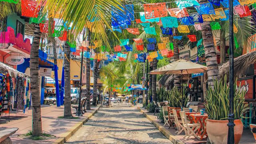 Colorful papel picado banners hanging above a palm-lined cobblestone street with shops and outdoor cafés in Sayulita, Riviera Nayarit, Mexico.